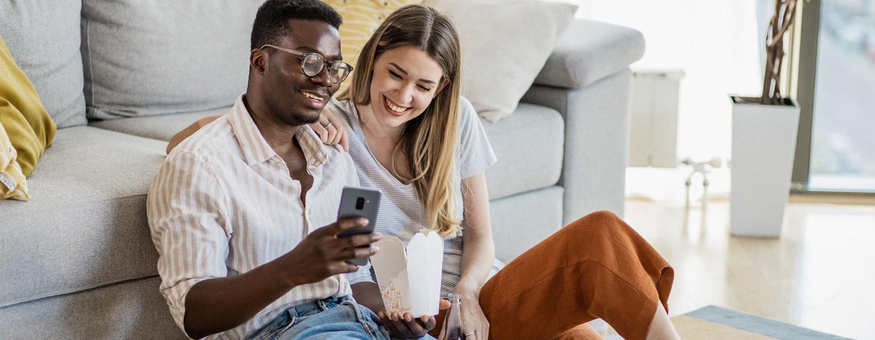 a man and woman with take out, sit against a couch on their living room floor and watch at their cell phone